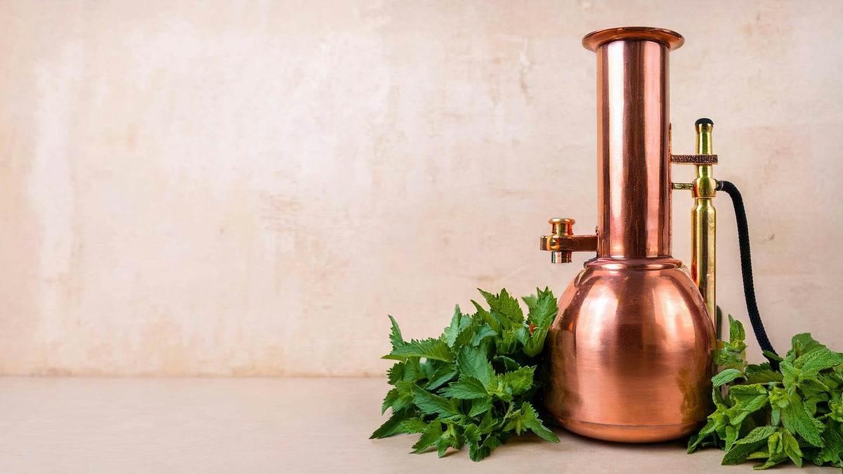 A copper distillation still beside fresh mint sprigs on a soft cream surface — botanical extraction in process.