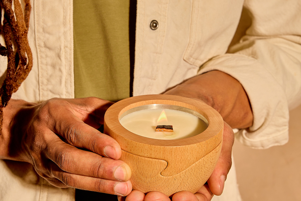 A person holds a lit Sero candle in its beechwood container, with the wood wick visible and a steady, clean flame.
