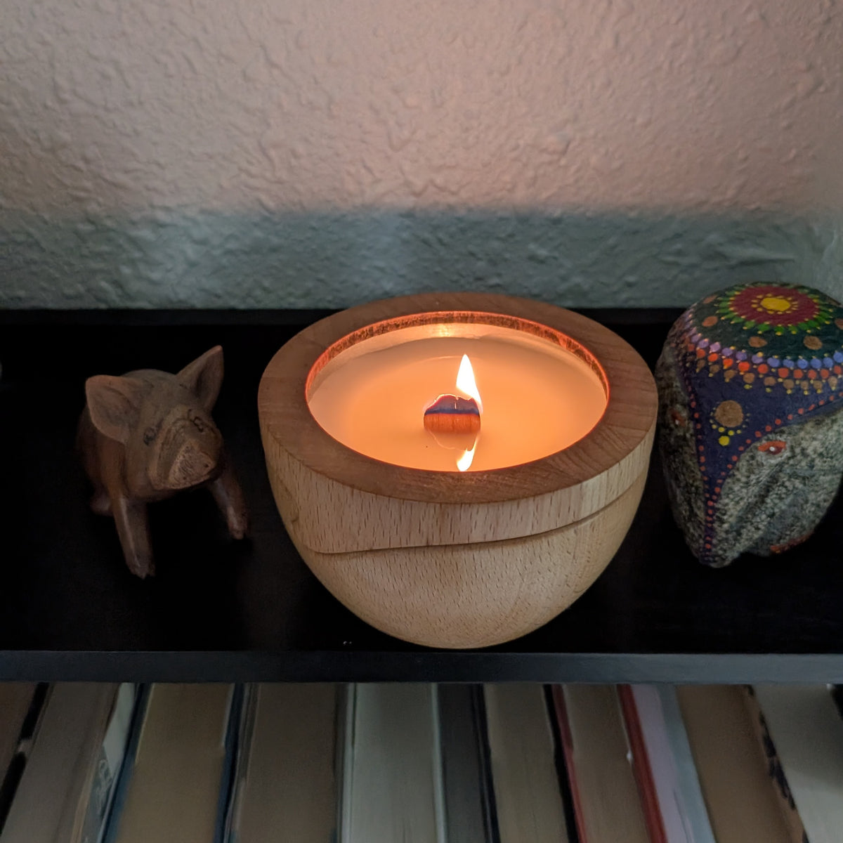 A lit candle in its wood container on a dark shelf above a row of books, the flame small and steady