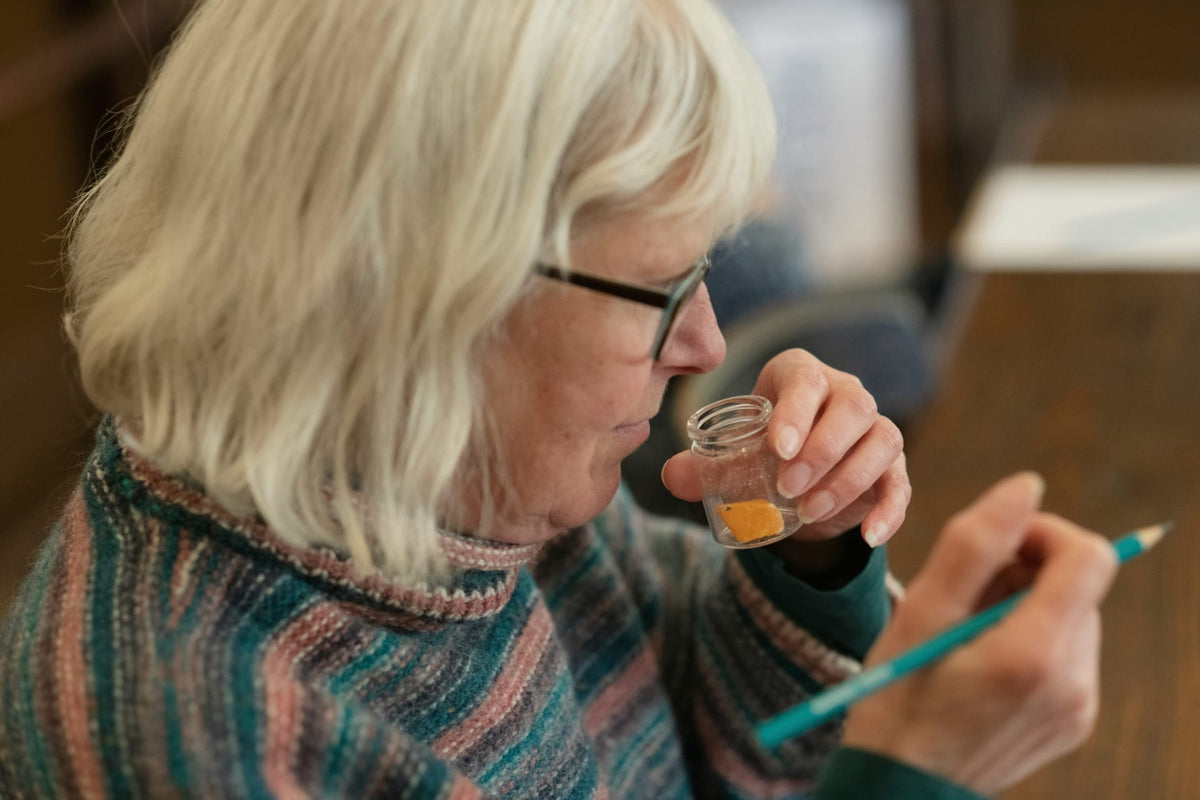 An older woman in profile holding a small glass fragrance vial up to her nose, evaluating its scent — a perfumer at work