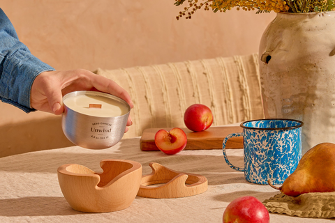 Person holding a candle labeled 'Unwind' on a table with apples, a mug, and decorative elements.