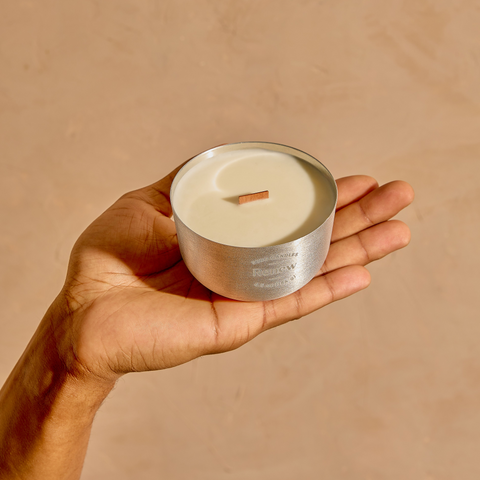 Hand holding a small silver candle container with a lit candle against a beige background